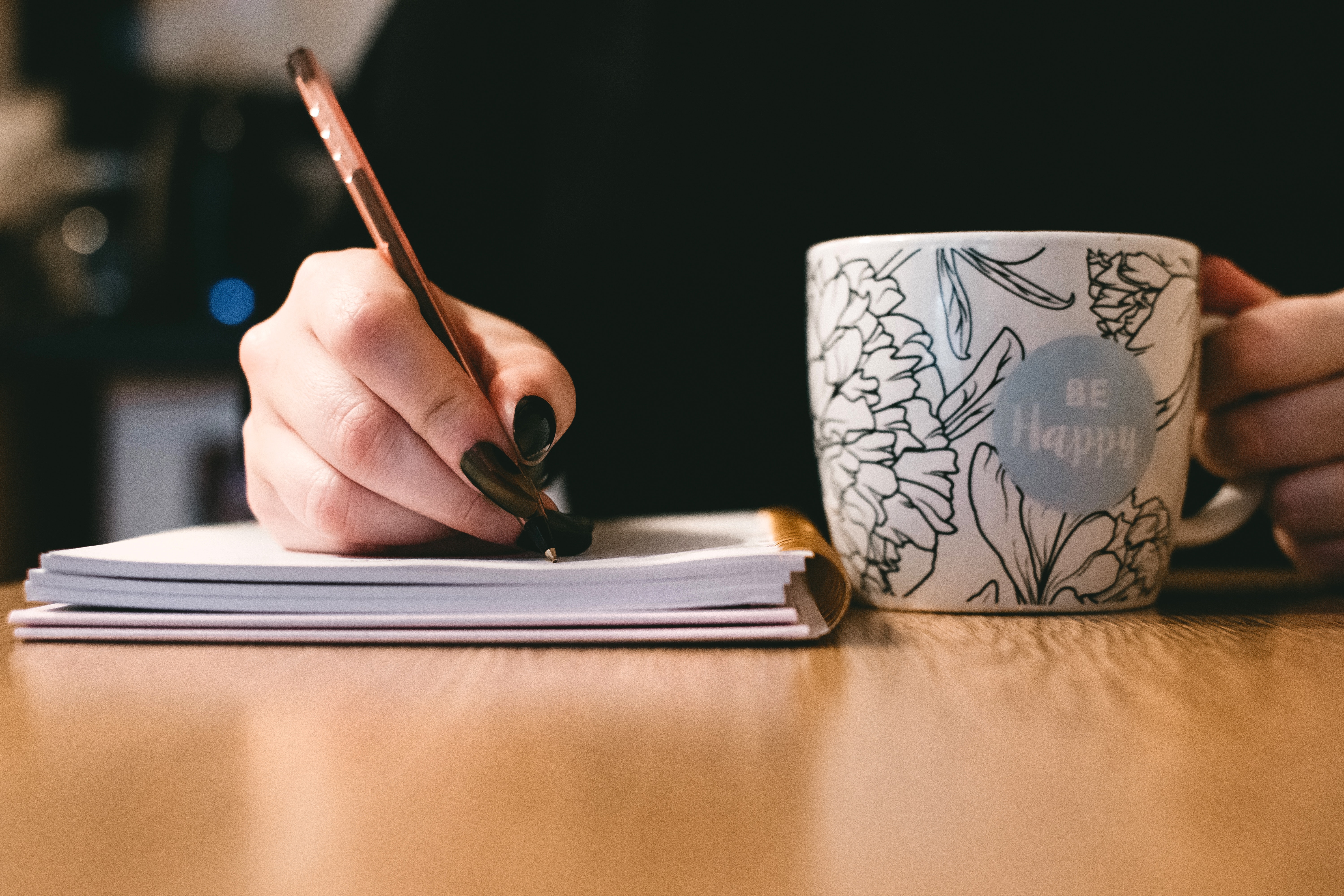 A person writing in a notebook and holding a mug
