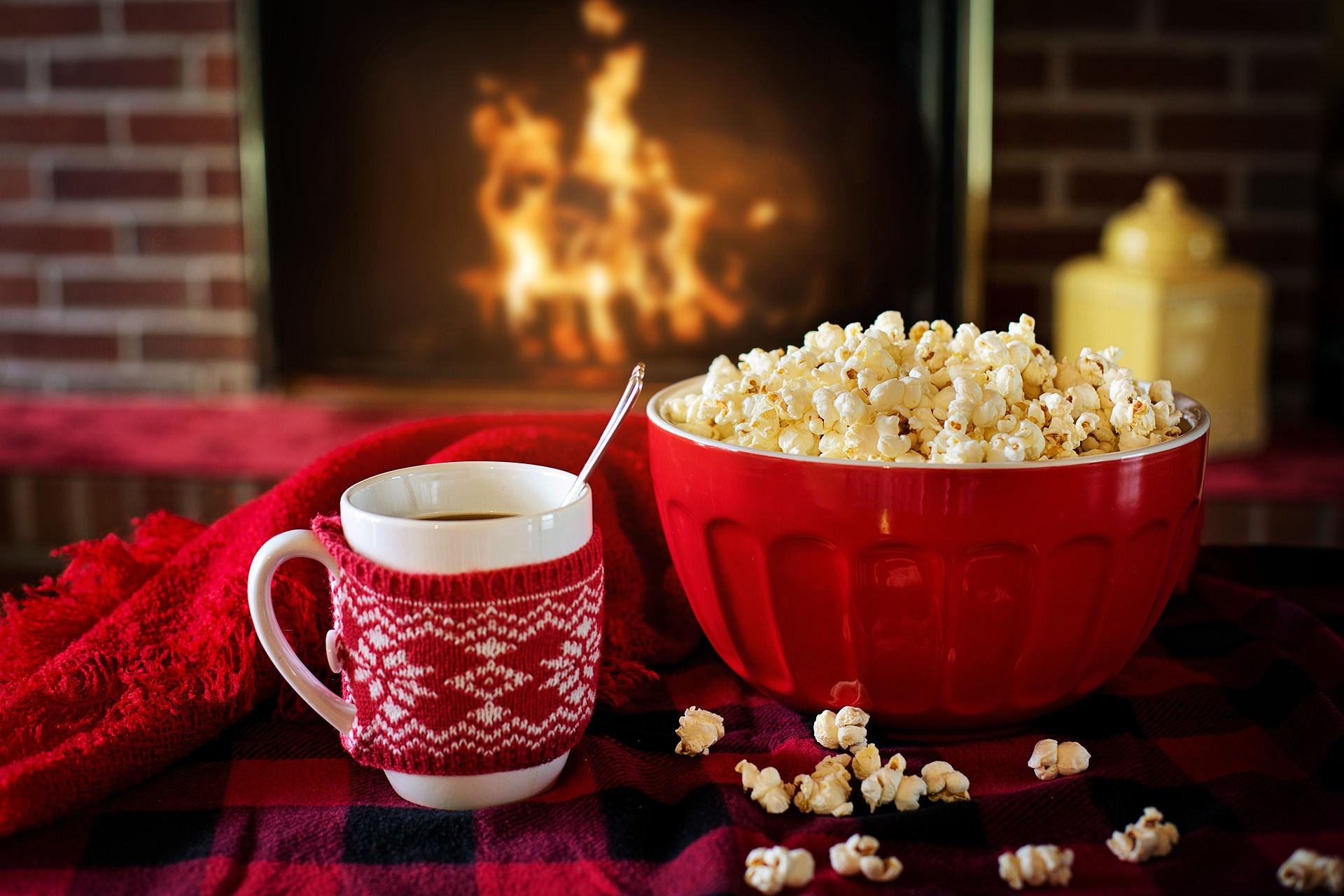 A bowl of popcorn next to a mug of hot cocoa both in front of a fire in a fireplace